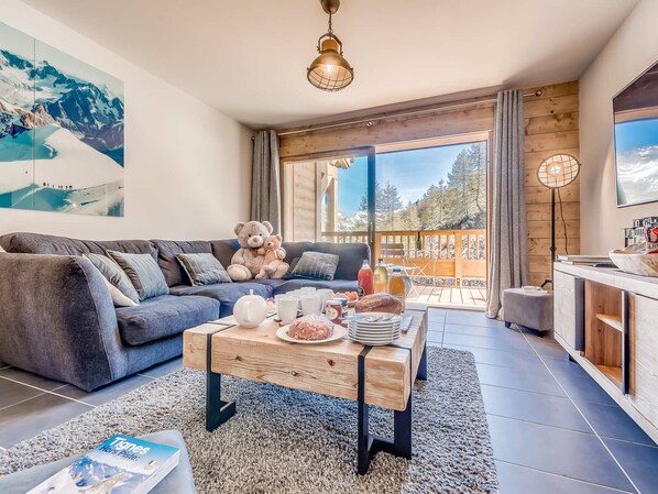 Ein modernes Wohnzimmer mit einer Couch, einem Couchtisch mit Getränken und Snacks, einem Teddybären und einem großen Fenster mit Bergblick.