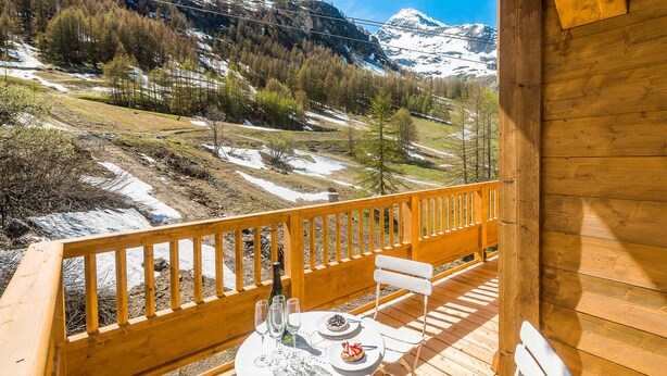 A wooden deck with a table and chairs, overlooking a mountainous landscape with snow patches and trees.