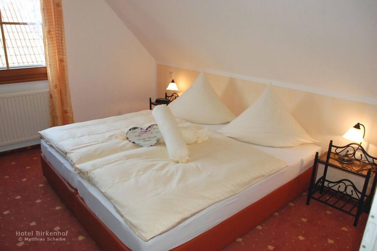 Uma cama de solteiro com lençóis e almofadas brancas em um quarto com teto inclinado. Há duas mesas de cabeceira com luminárias.