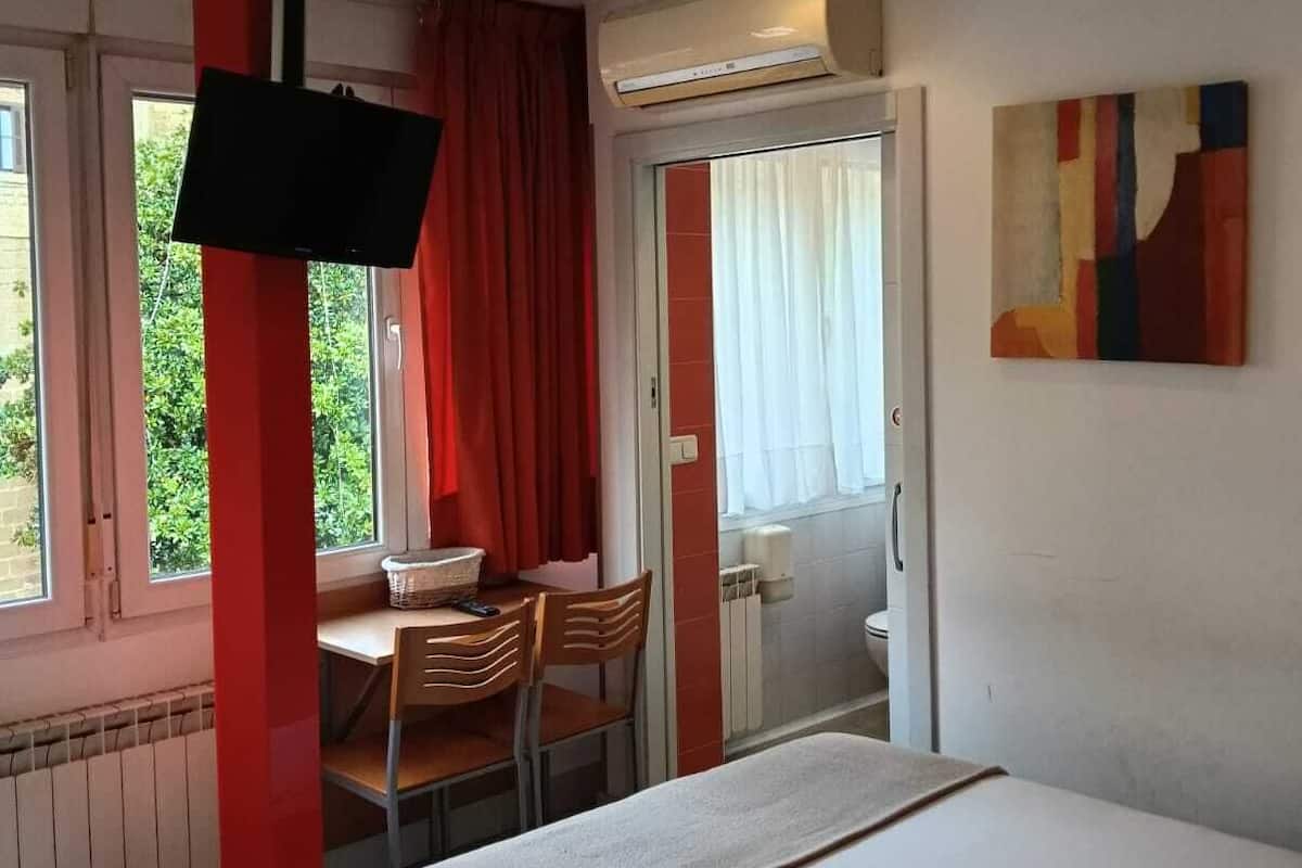 A bedroom with a bed, a desk with a chair, a television, and a window with curtains.