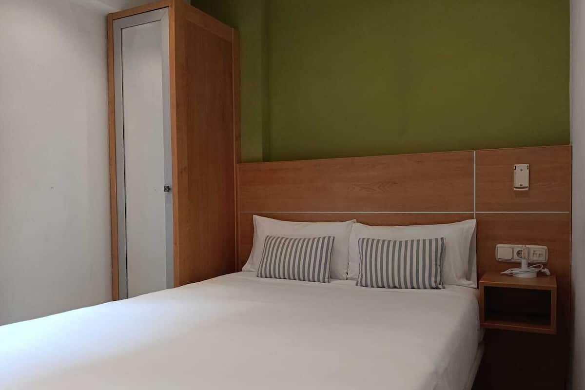 A single bed with a wooden headboard, a bedside table with a lamp, and a power outlet on the wall.