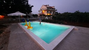 A rectangular swimming pool with a tiled edge, surrounded by a paved area. There are two umbrellas labeled "PAN" and a building with a balcony in the background. - NEW Apartment, A2+2, Family house - OPG Rudan, garden pool, terase, wi-fi (Drnis)