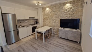 A modern kitchen with white cabinets, a stainless steel refrigerator, a stone accent wall, a dining area with a table and chairs, and a flat-screen TV on a grey cabinet. - NEW Apartment, A2+2, Family house - OPG Rudan, garden pool, terase, wi-fi (Drnis)
