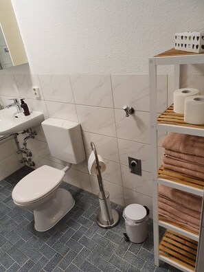 A bathroom with a toilet, sink, and shelves with towels. - 2 Ferienwohnungen in Schweinfurt (Schweinfurt)