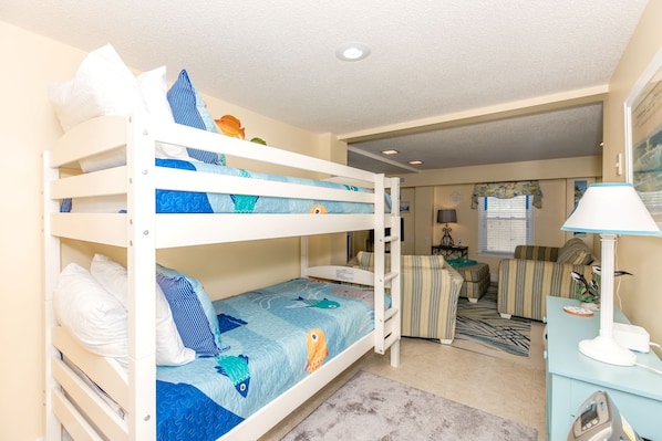 A bunk bed with a blue and white patterned bedspread, a small desk with a lamp, and a striped sofa in a room with beige walls and a ceiling light.