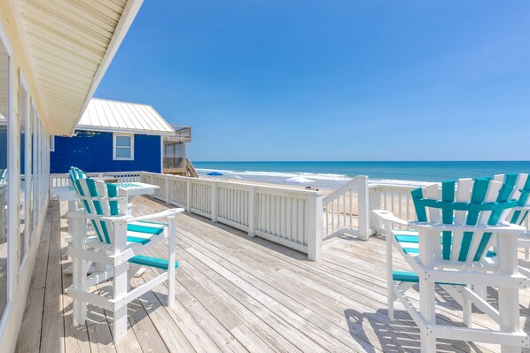 A wooden deck with white and teal Adirondack chairs, a small blue building, and a view of the ocean.