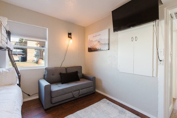 A cozy room with a grey sofa, a white radiator, a mounted TV, and a window with blinds.