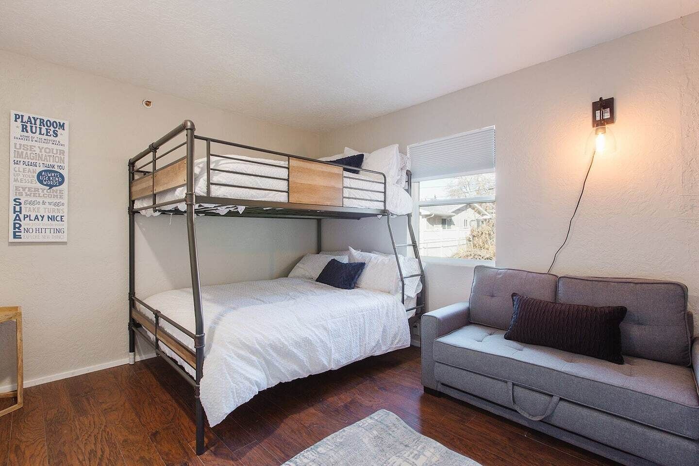 A bunk bed with a playroom rules sign, a grey sofa, and a window with blinds.