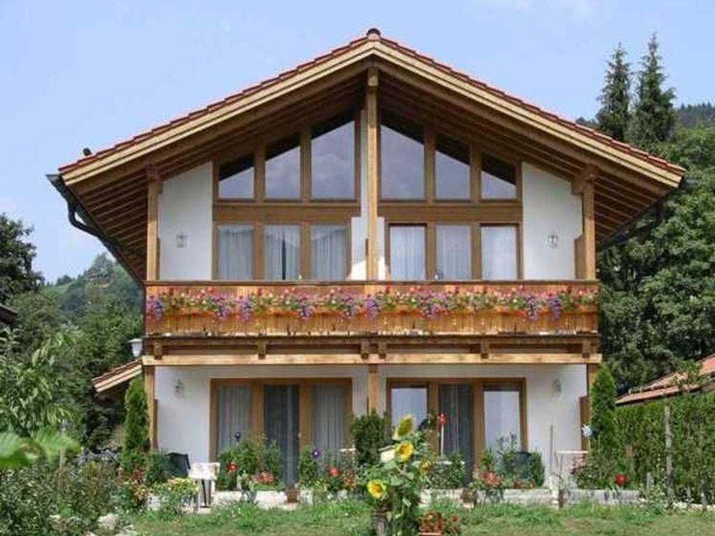 Un chalet à deux étages doté d’un balcon en bois, de grandes fenêtres et d’un jardin à l’avant.