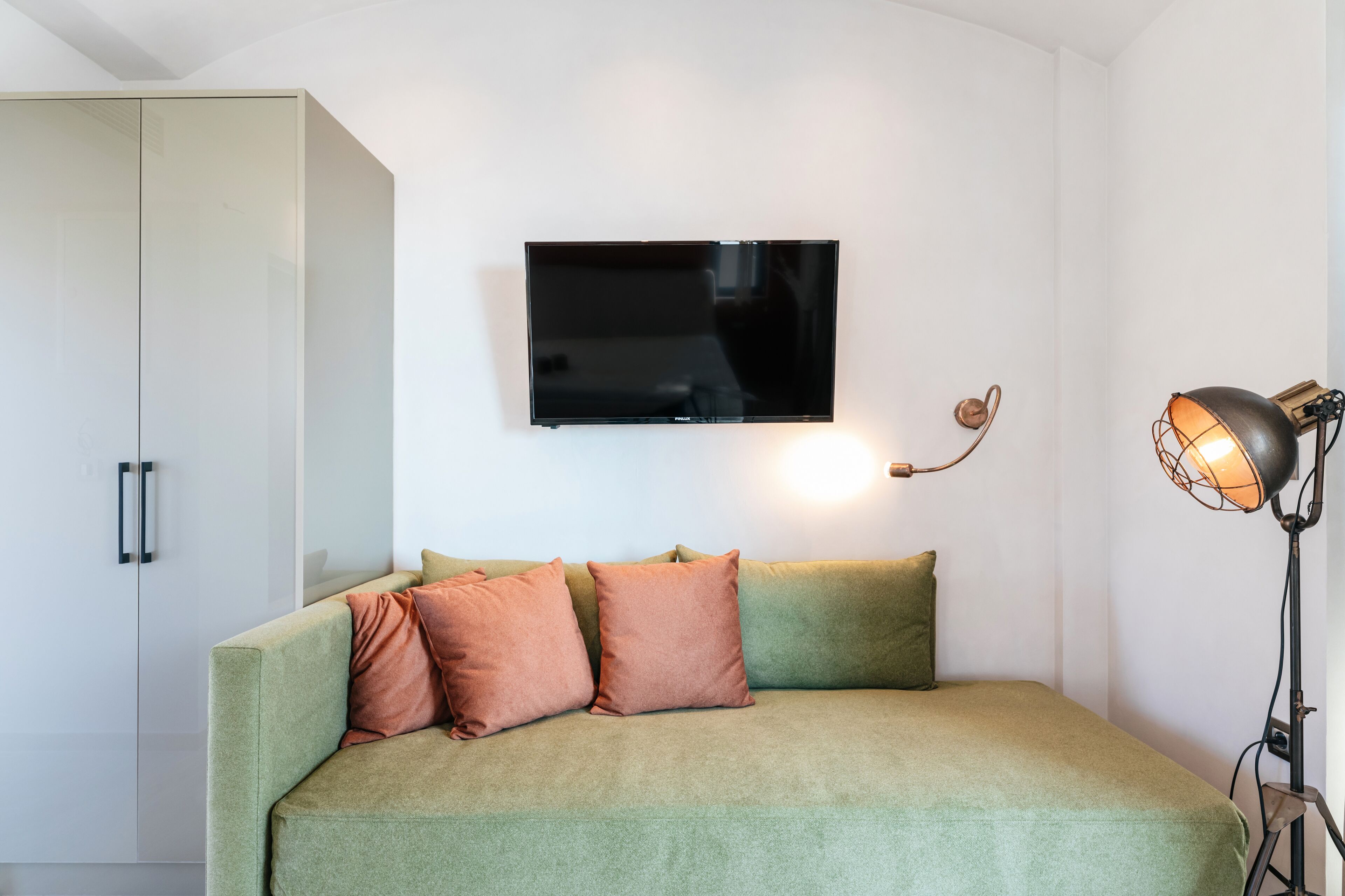 Een moderne slaapkamer met een groen slaapbankje, een flatscreen televisie aan de muur en een vloerlamp in vintage-stijl.