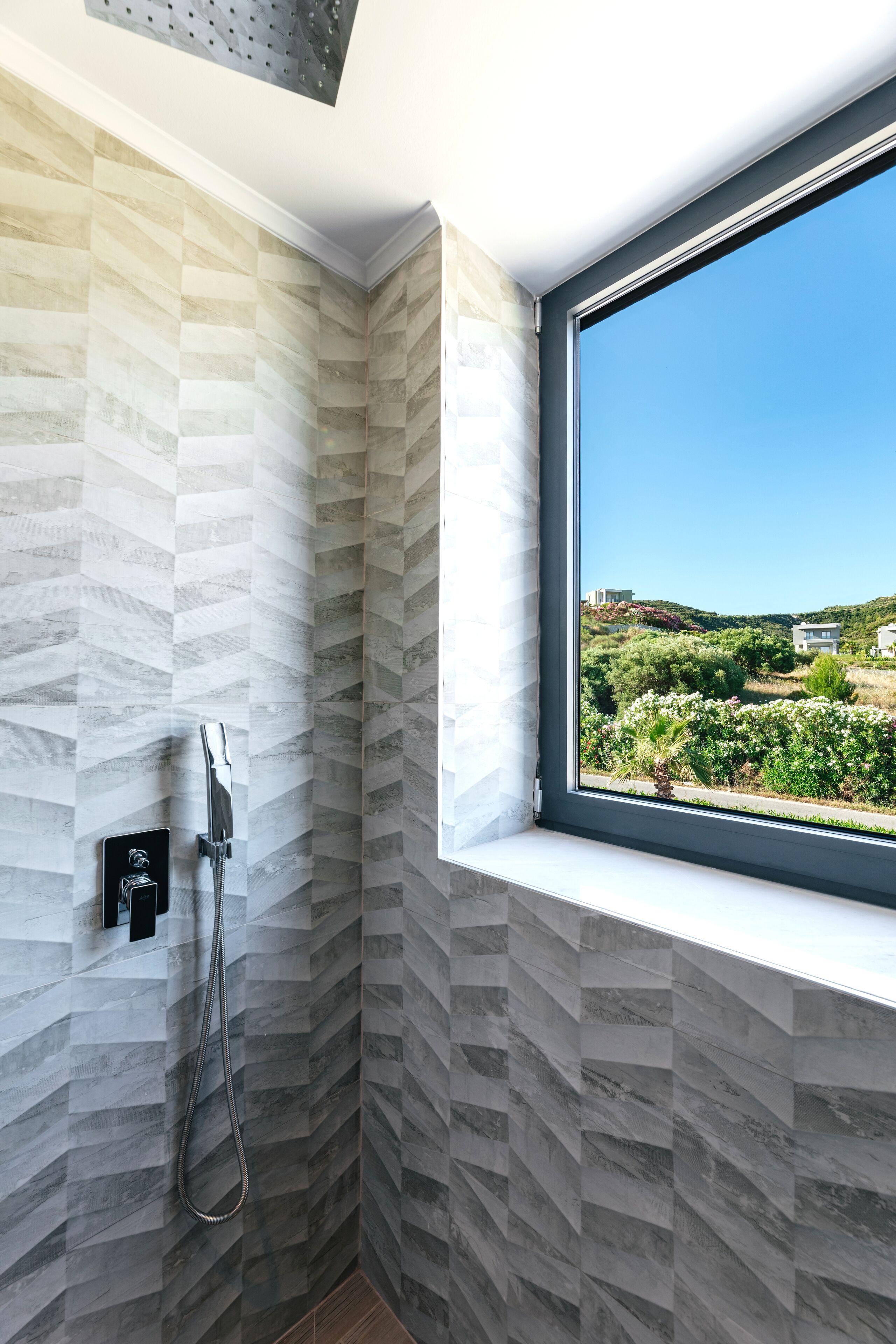 A bathroom with a shower, a window with a view of greenery and buildings, and a wall with a geometric pattern.