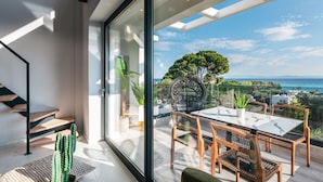 Un interior moderno con un amplio ventanal que da a una terraza exterior con mesa y sillas, un cactus y una escalera.