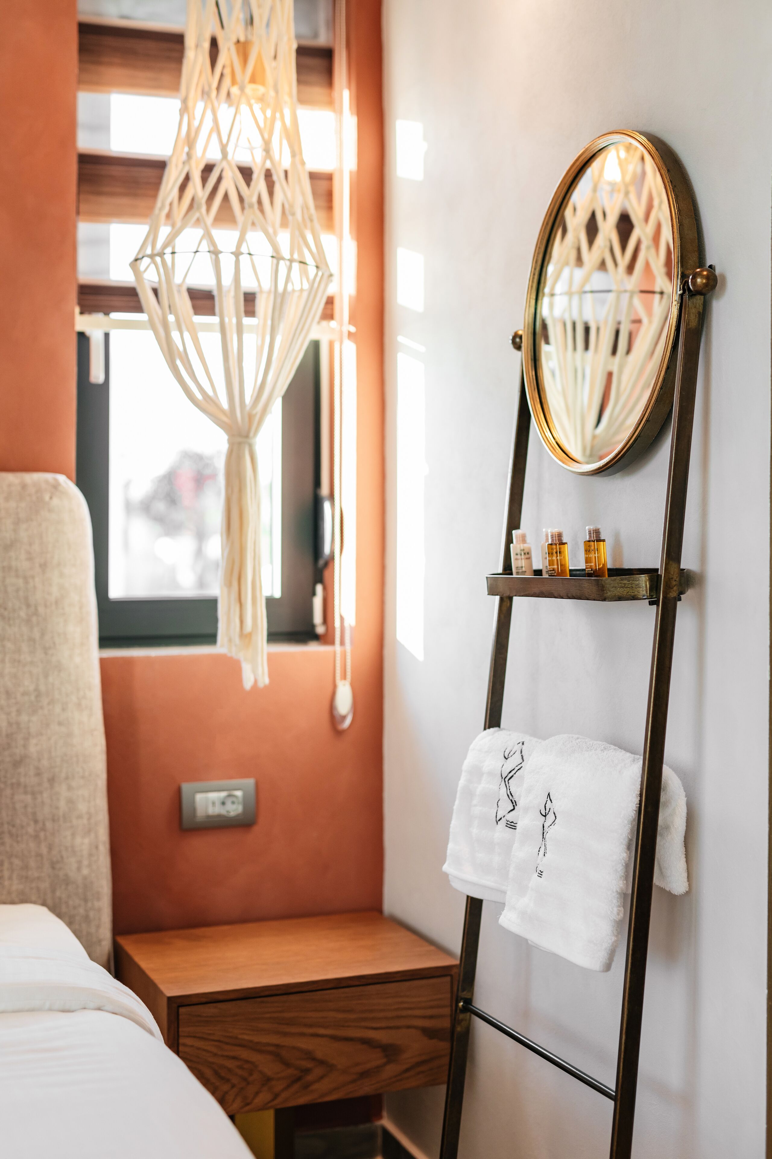 Una habitación de hotel con una cama, una mesita de noche de madera, un estante con artículos de tocador y una lámpara colgante de macramé, además de un espejo con una toalla colgada.