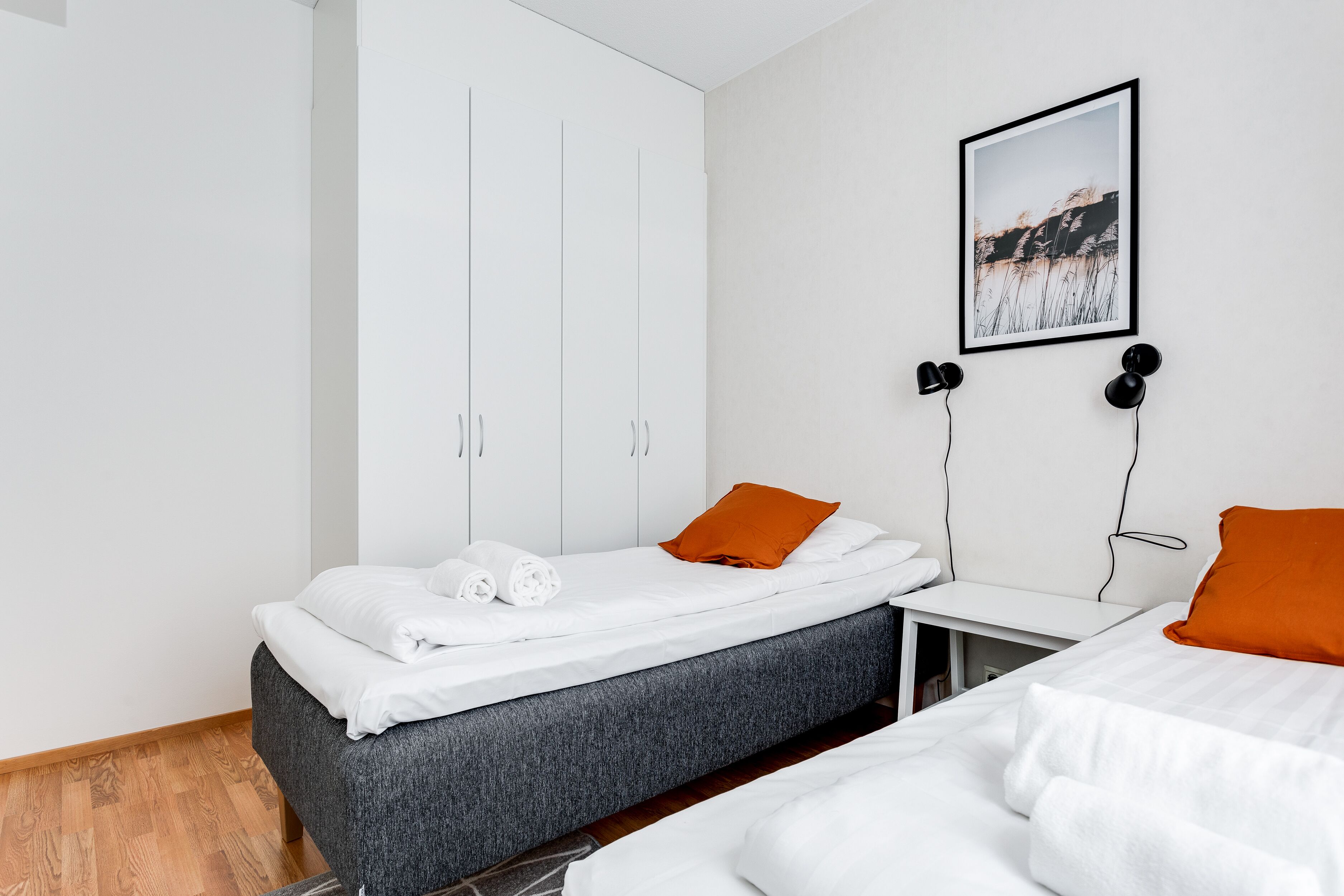 Une chambre moderne avec un lit simple, une tête de lit grise, du linge de lit blanc et un coussin orange. On y trouve une armoire blanche avec un miroir et un tableau encadré accroché au mur.