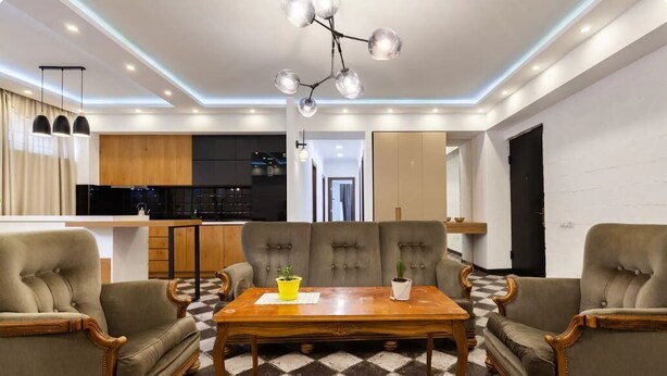 Un salon moderne avec une table basse en bois, deux fauteuils et un canapé. Un lustre est suspendu au-dessus de la table, et on aperçoit une cuisine au fond.