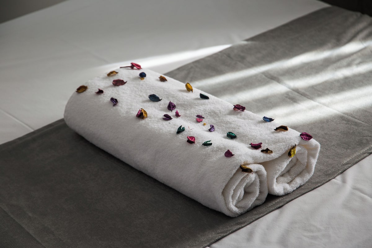 A neatly folded white towel with colorful flower petals on a bed with a grey bedspread.