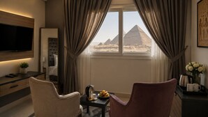 Habitación de hotel con televisor, cama, mesa de comedor con fruta y vista a pirámides.