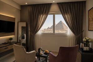 A hotel room with a TV, a bed, a dining table with fruit, and a view of pyramids. - Gardenia Pyramids View Inn - Rooftop (Giza)