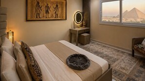 Un dormitorio moderno con una cama grande, una obra de arte en la pared, una tocador con espejo y vistas a pirámides a través de un ventanal.