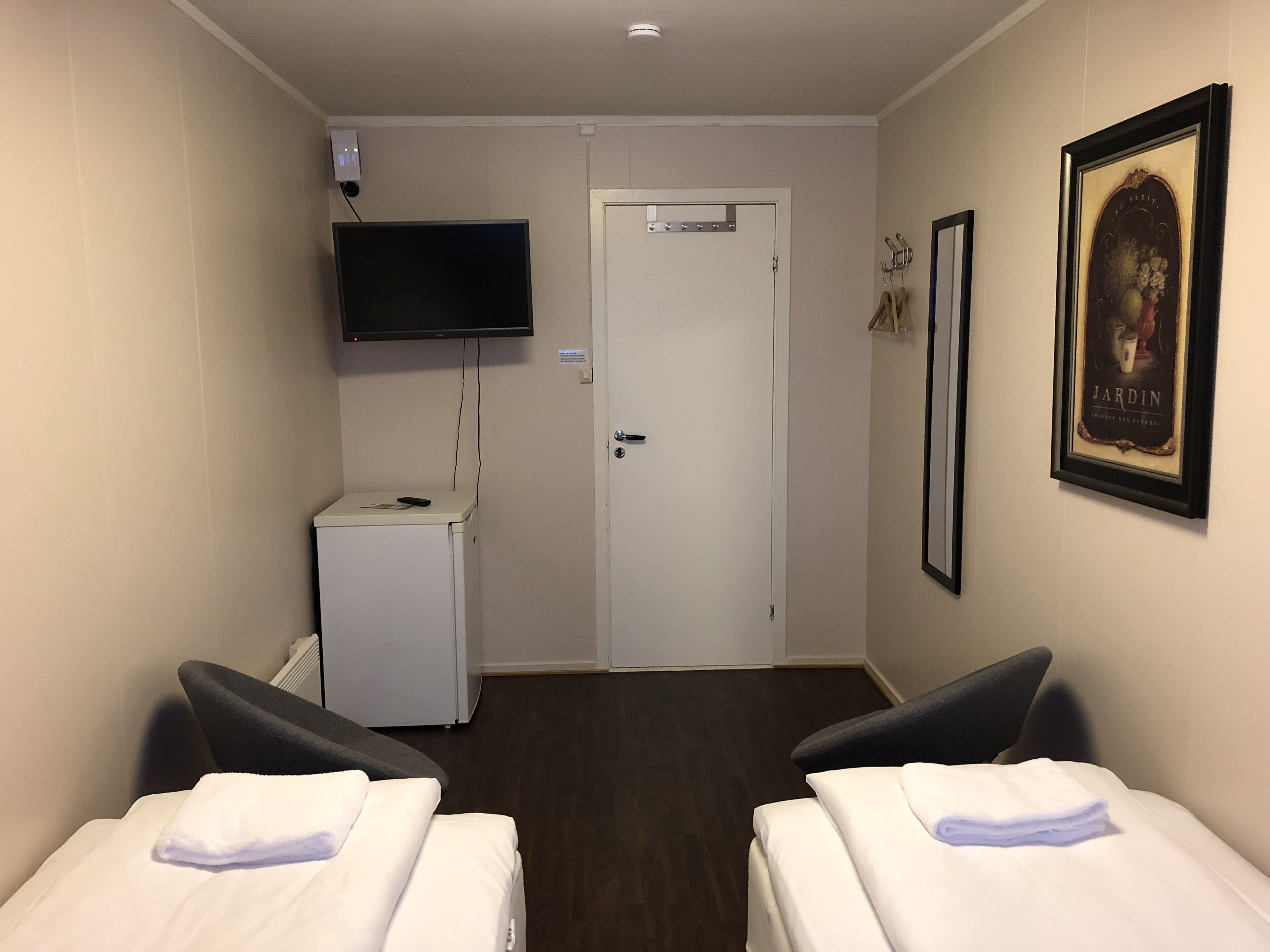 A hotel room with two beds, a television mounted on the wall, a small white cabinet, and a door with a sign above it.
