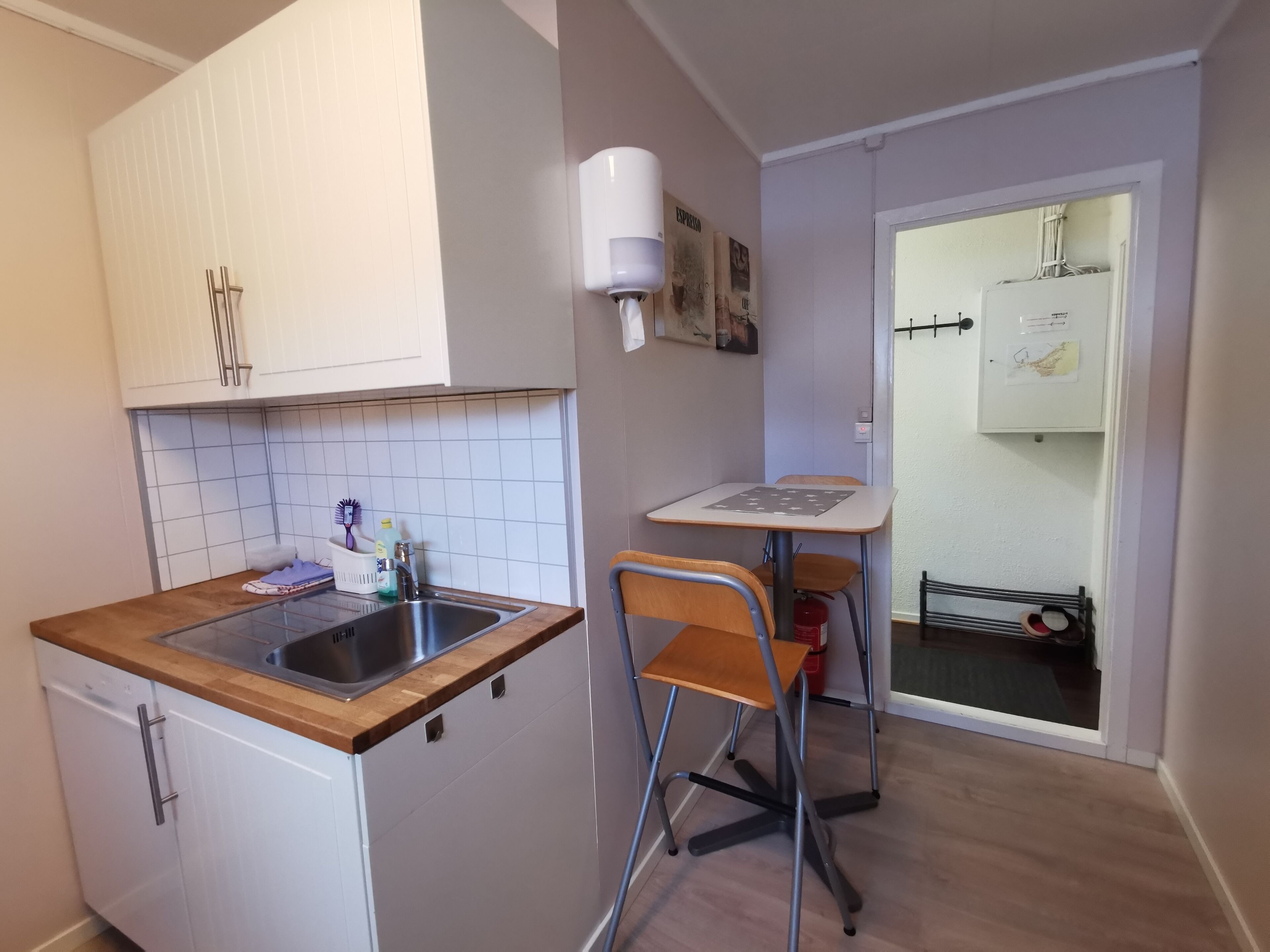 A compact kitchen with a sink, a small table, and a chair. There is a white cabinet with a handle, a tiled backsplash, and a white wall with a light switch.