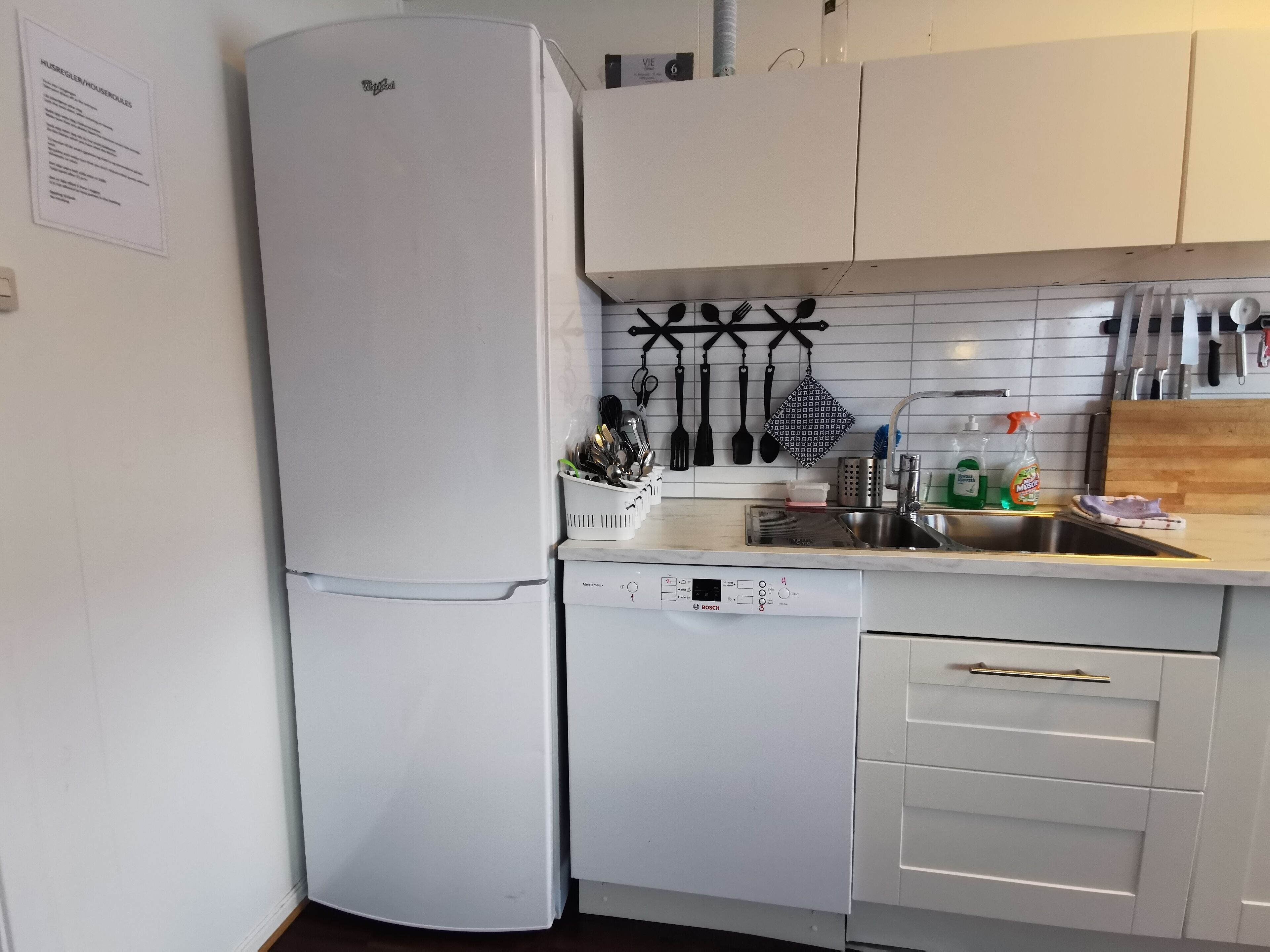 Una cocina con armarios blancos, un lavavajillas Bosch y un refrigerador. Hay utensilios colgados en la pared y un fregadero con tabla de cortar.