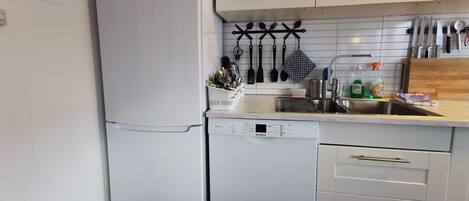 Una cocina con armarios blancos, un lavavajillas Bosch y un refrigerador. Hay utensilios colgados en la pared y un fregadero con tabla de cortar.