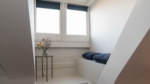 Une petite pièce bien éclairée, avec une fenêtre, une petite table sur laquelle est posé un vase de fleurs et un lit avec des oreillers.