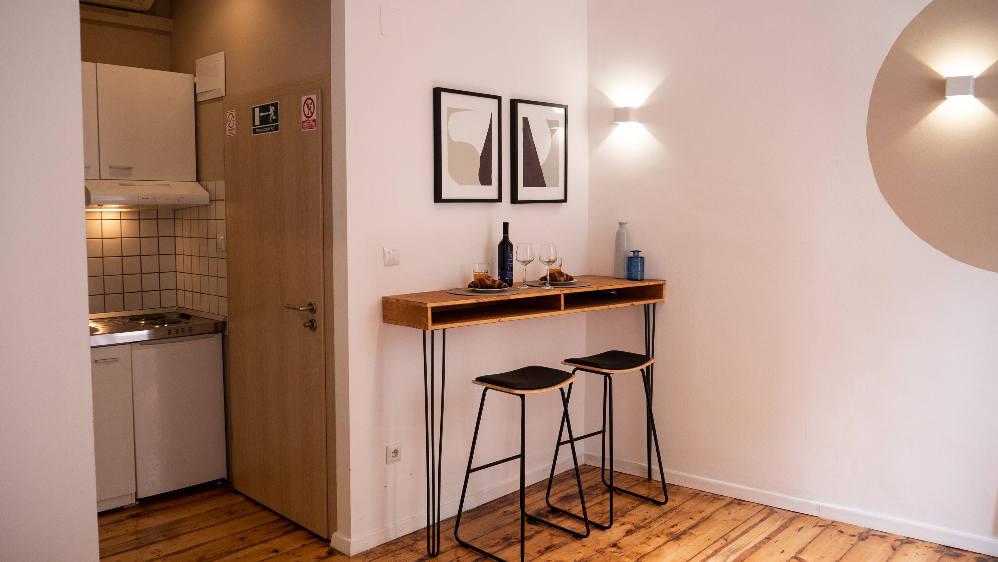 A compact kitchen with a wooden door, a small dining table with two stools, and wall-mounted artwork.
