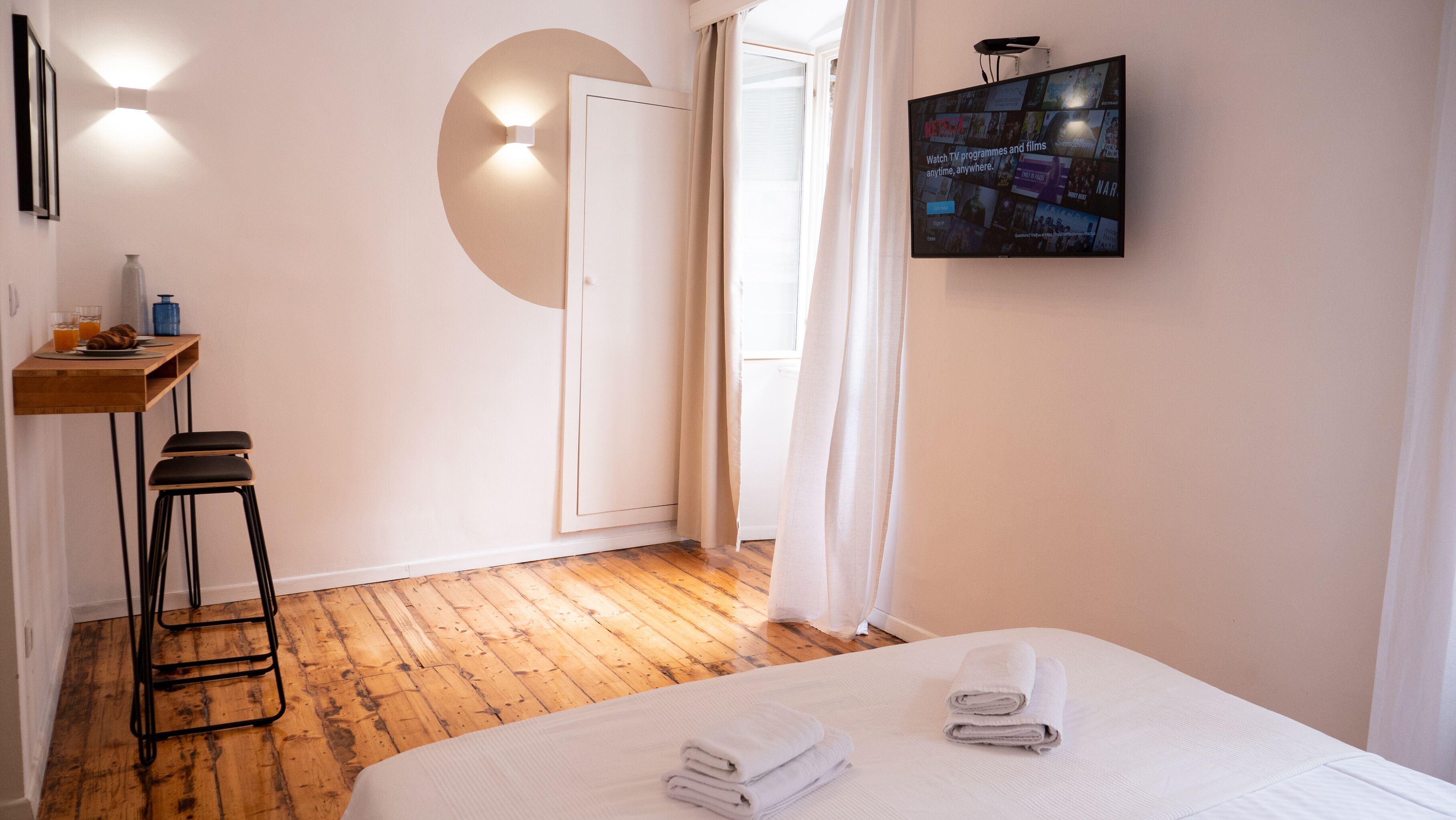 A hotel room with a wooden floor, a bed with white linens, a small desk with a chair, a wall-mounted TV, and a door with curtains.