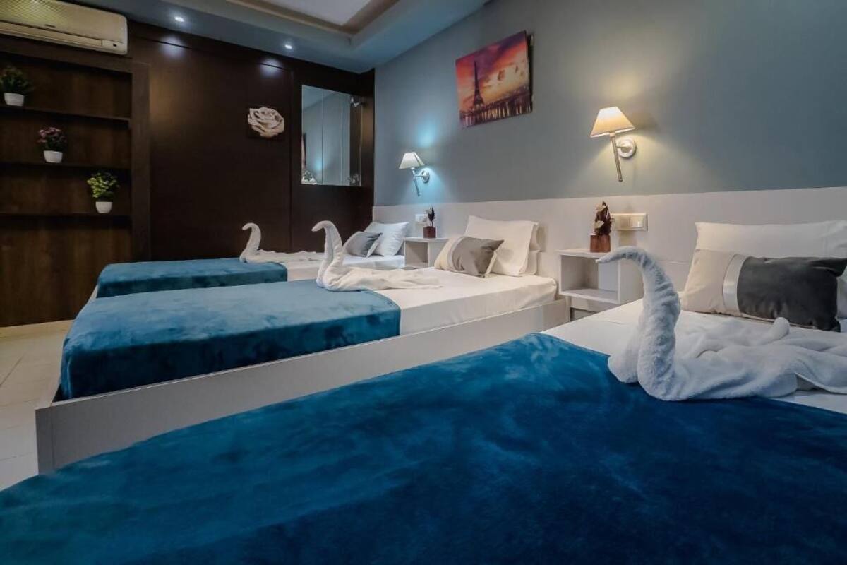 Une chambre d’hôtel avec deux lits, chacun recouvert d’une couvre-lit bleue et agrémenté d’oreillers blancs, et un cygne décoratif posé sur chaque lit. On y trouve une table de chevet avec une lampe, un miroir fixé au mur et une étagère avec des plantes.