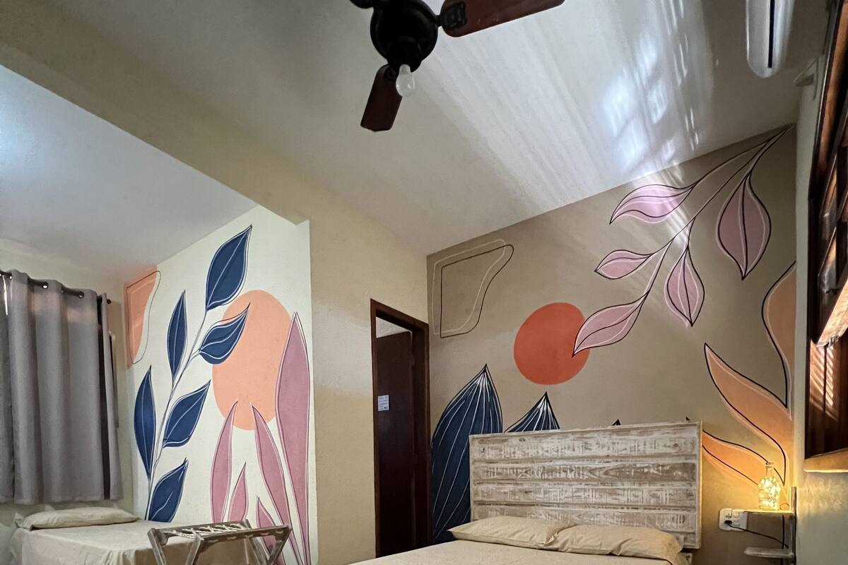 A hotel room with two beds, a ceiling fan, a wall with abstract art, and a bedside table with a lamp.