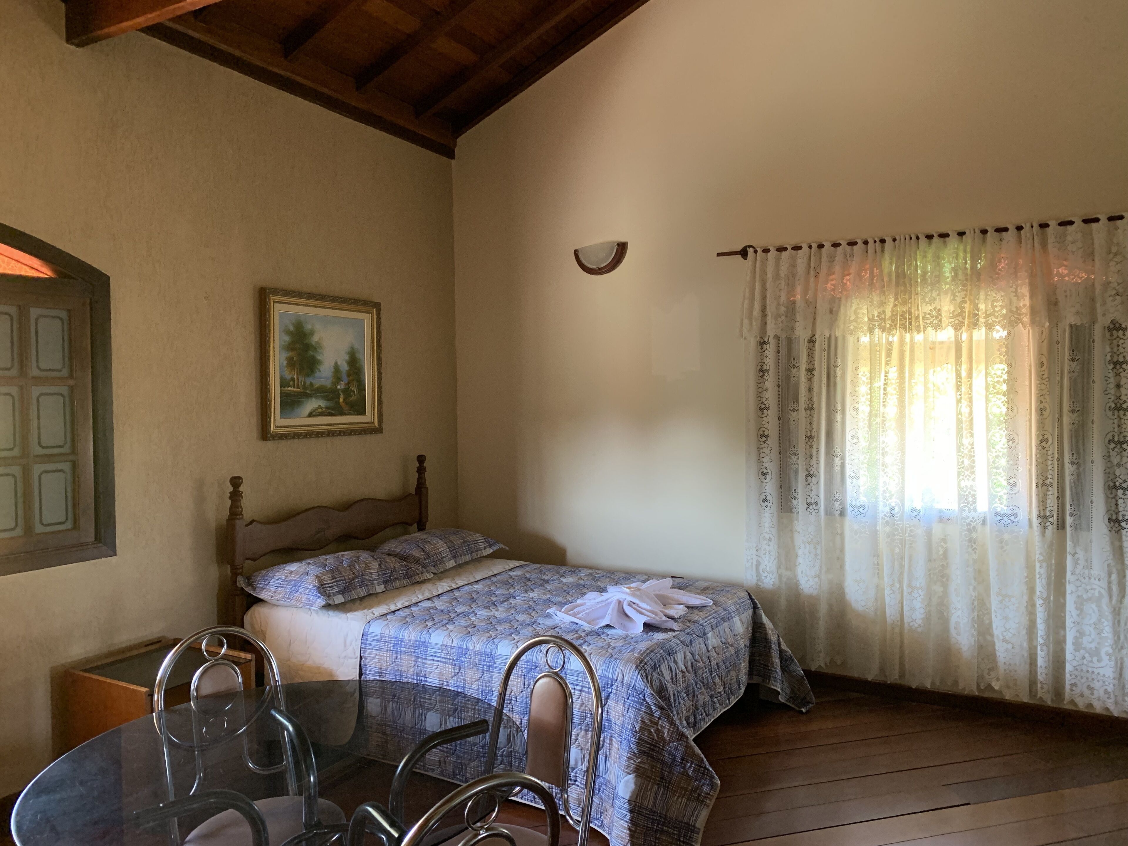 A bedroom with a wooden bed, a glass table with chairs, a painting on the wall, and a window with patterned curtains.