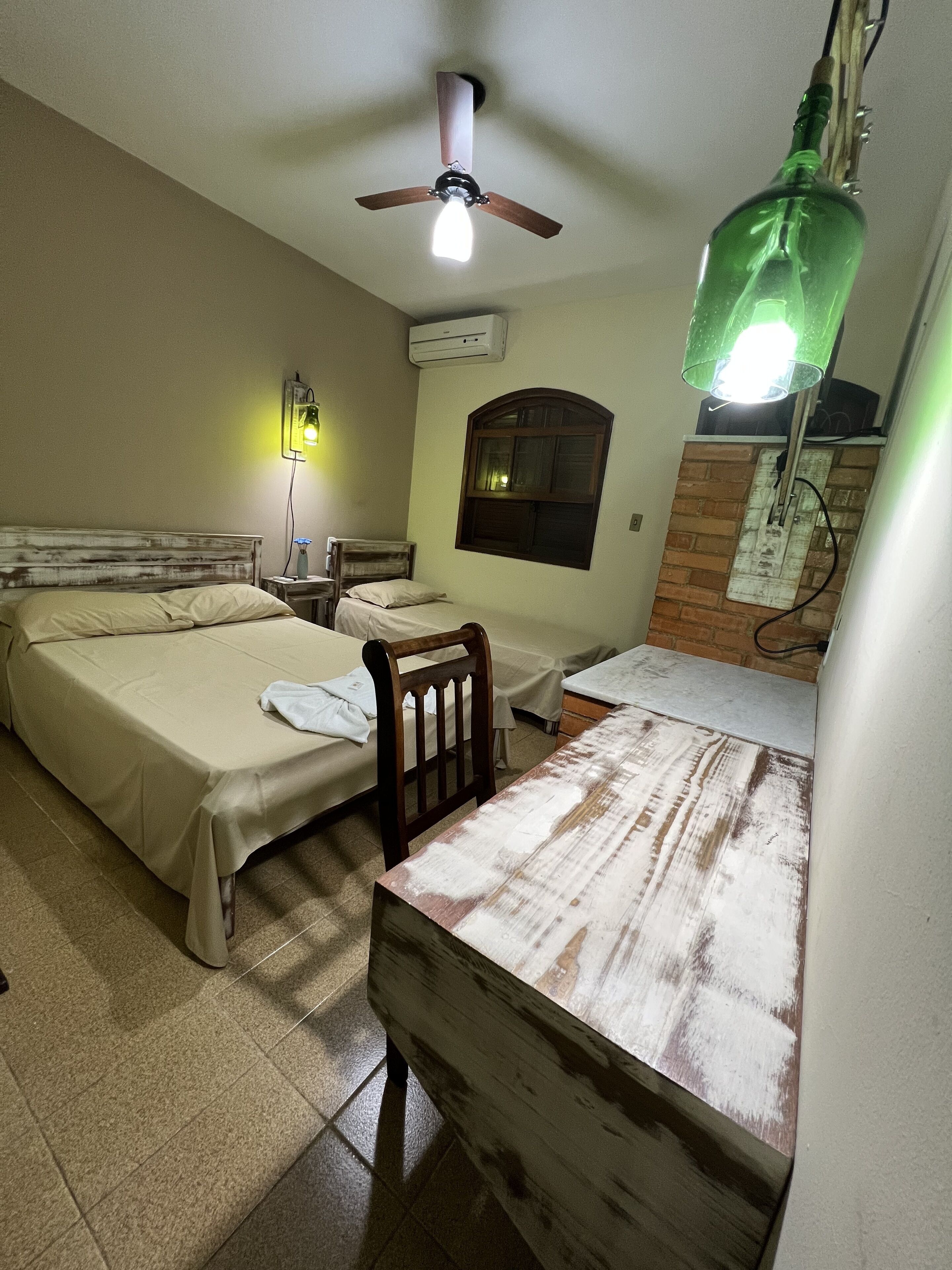 Quarto com duas camas, ventoinha no teto, ar condicionado, janela e uma mesa.