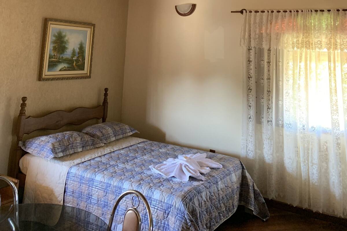 A bedroom with a wooden ceiling, a bed with a blue and white patterned bedspread, a painting on the wall, and a window with sheer curtains.