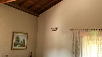 Un dormitorio con techo de madera, una cama con colcha de rayas azules y blancas, un cuadro en la pared y una ventana con cortinas de encaje.