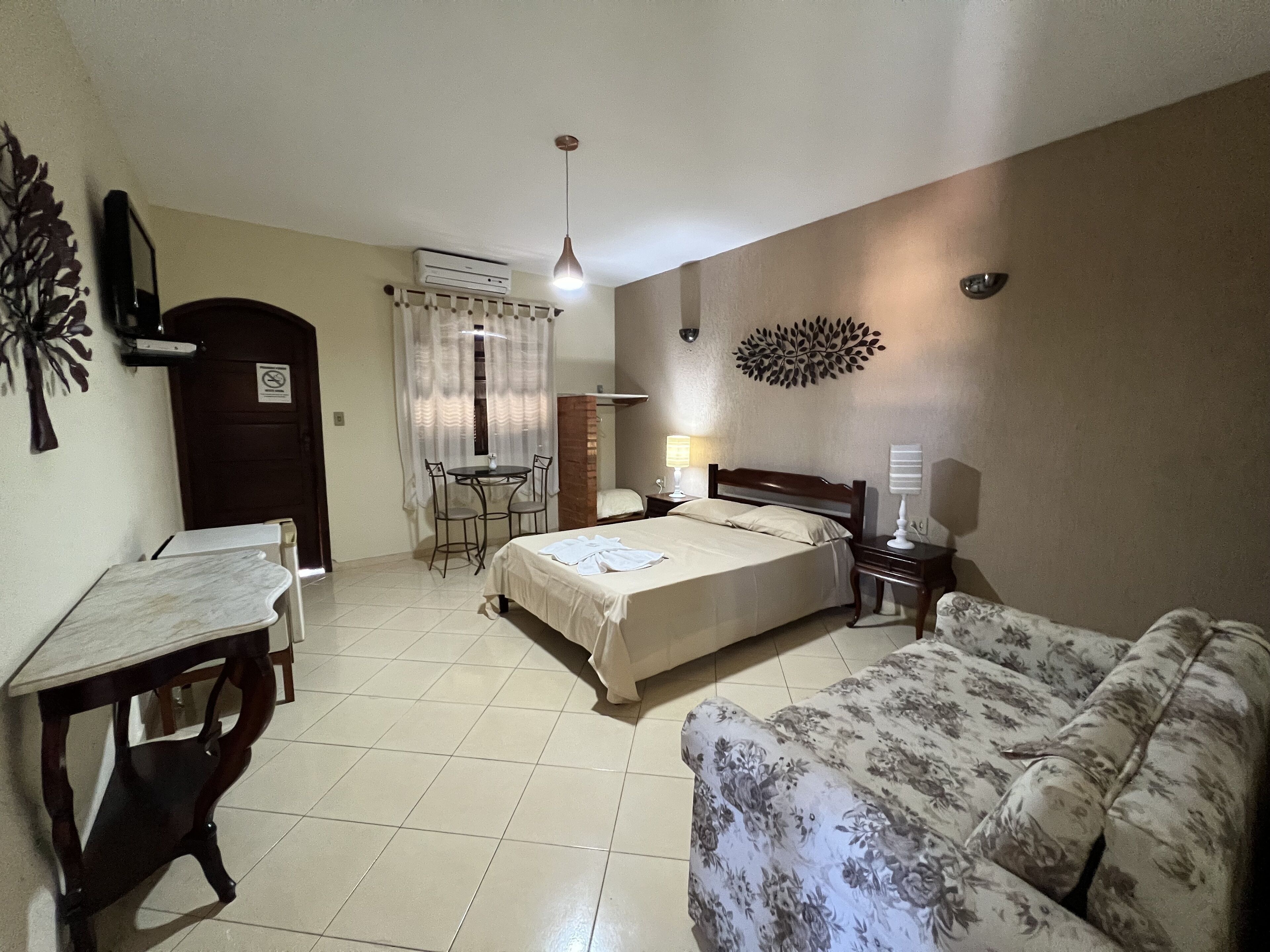 Quarto de hotel com cama, sofá, escrivaninha com cadeira, mesa pequena, luminária e um objeto decorativo na parede.