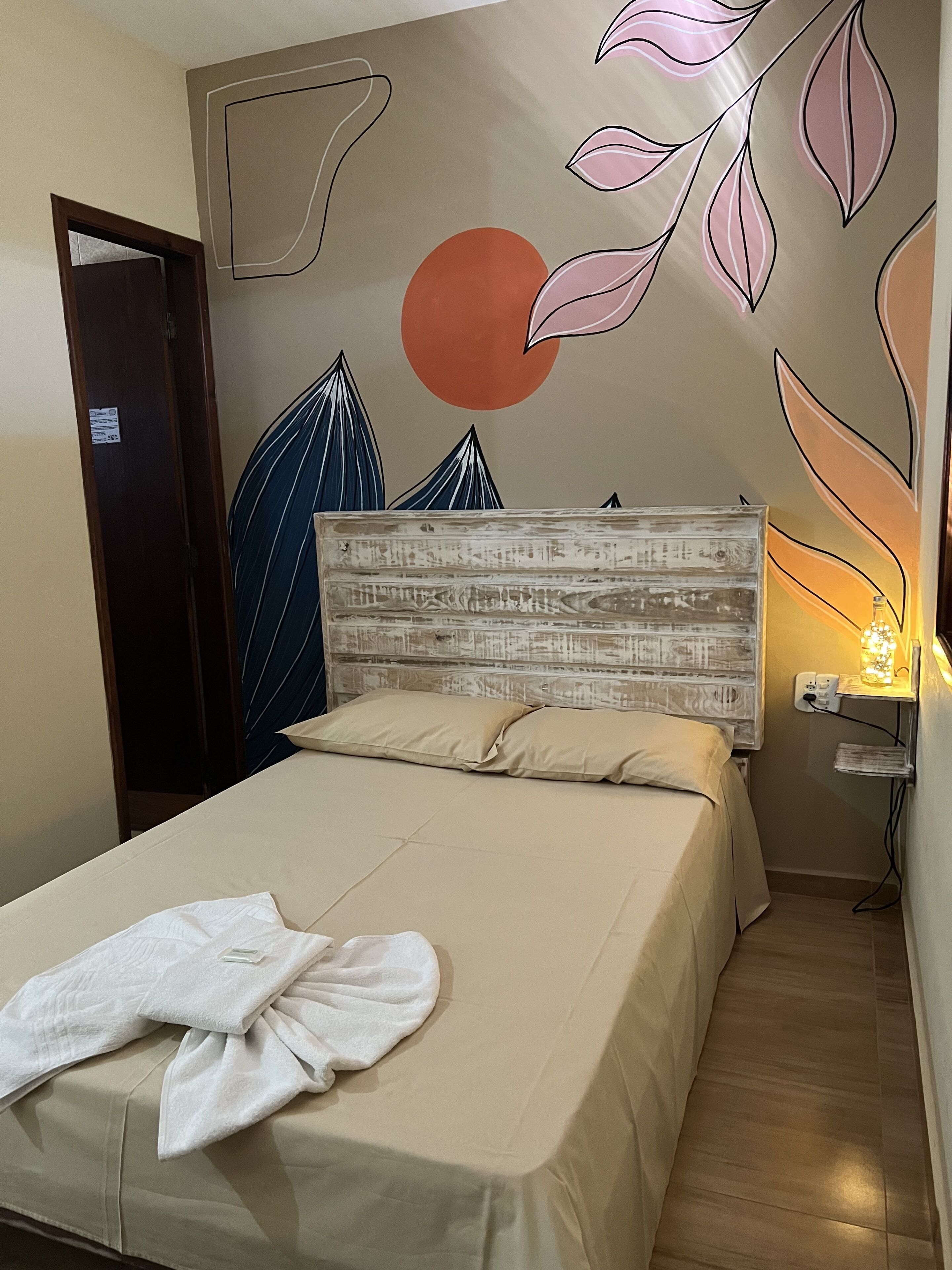 Una cama bien tendida con colcha y almohadas color beige, cabecera de madera y pared con diseño de hojas y motivo circular.