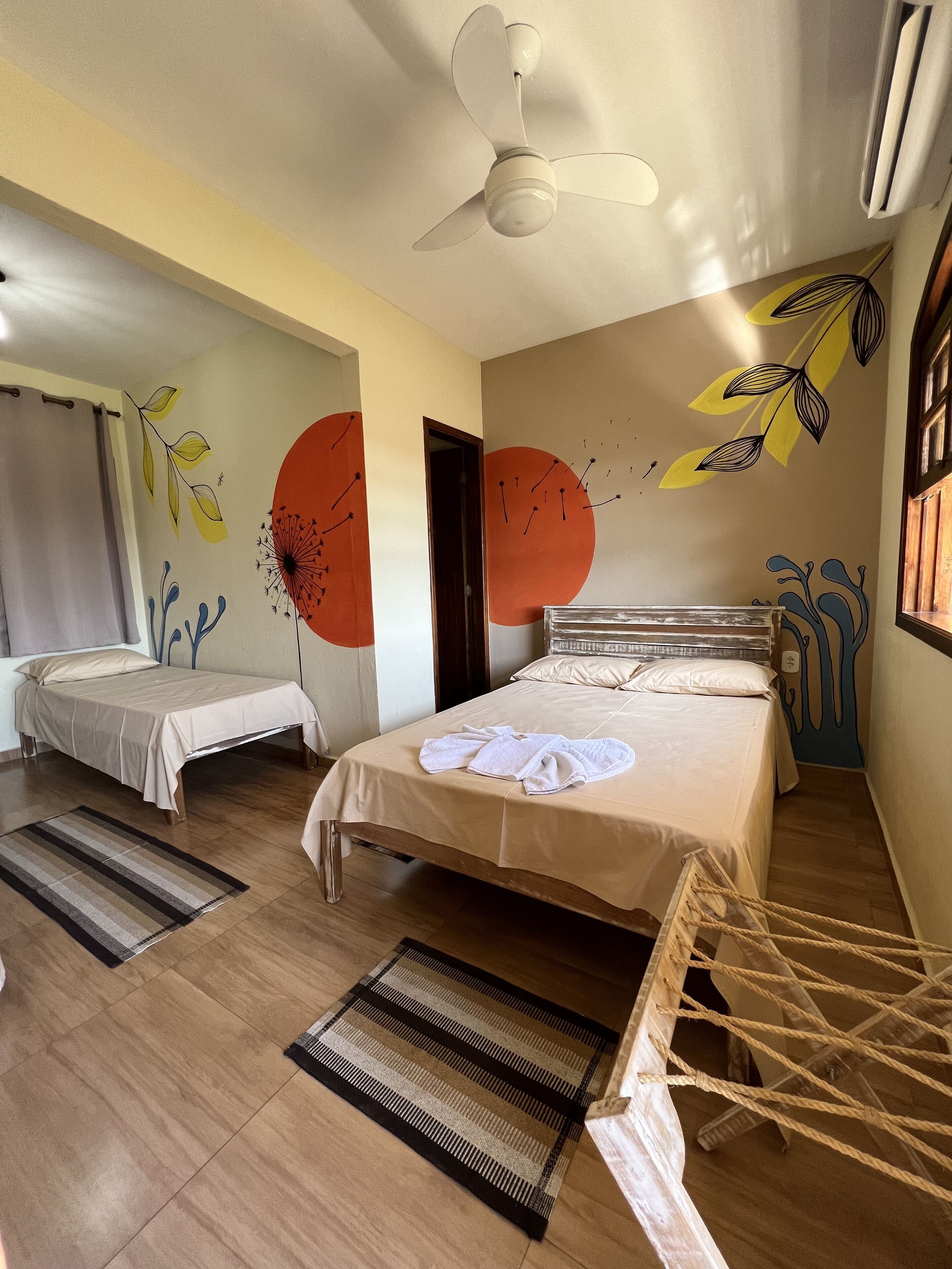 Habitació d'hotel amb dos llits, un ventilador de sostre, una finestra gran amb cortines i una paret decorada amb art abstracte.