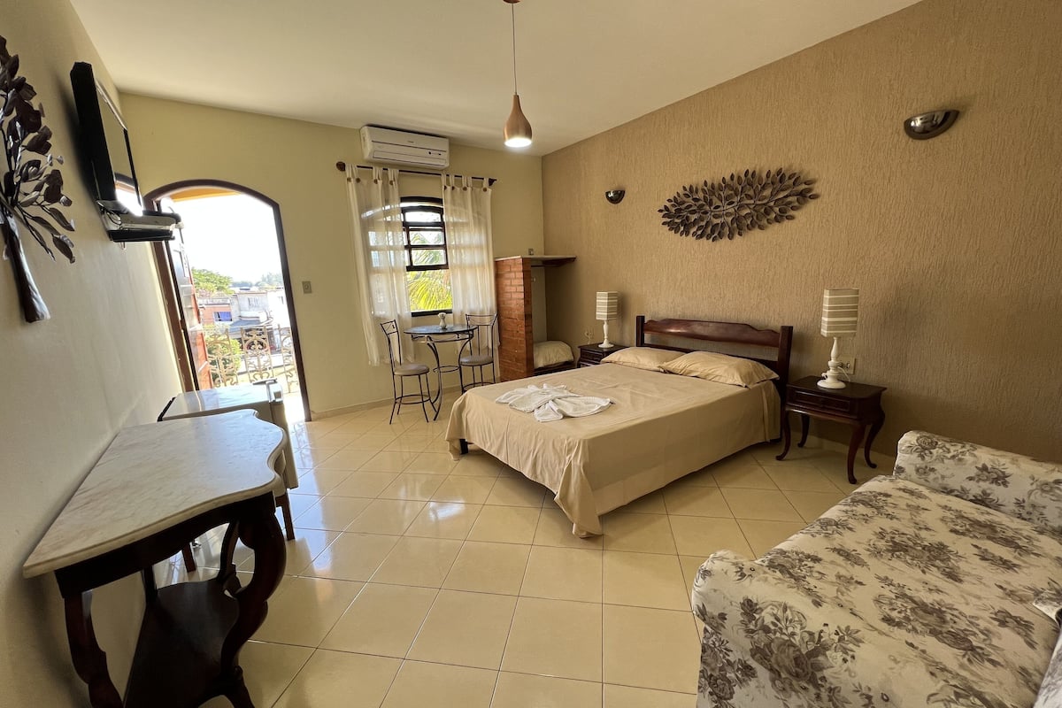 A hotel room with a bed, a desk, a chair, a small table, a lamp, and a decorative wall piece.