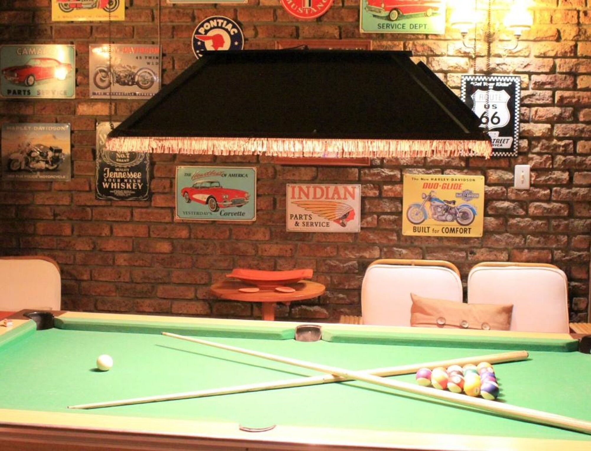 Una mesa de billar en una habitación con pared de ladrillo, varios letreros de servicios automotrices y dos sillones acolchados.
