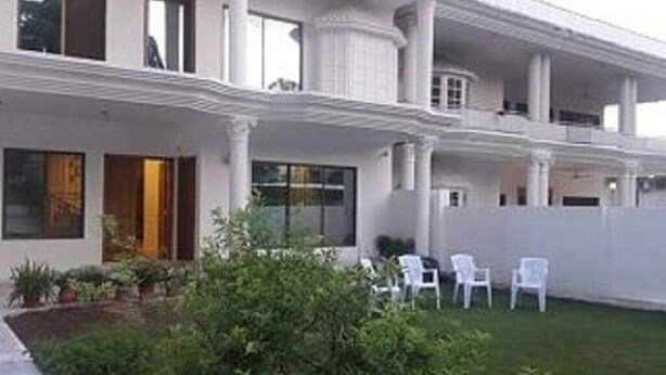 Una casa de dos pisos con un balcón, un jardín con plantas en macetas y sillas blancas.
