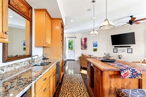A modern kitchen with granite countertops, wooden cabinets, a large island, pendant lights, and a ceiling fan.