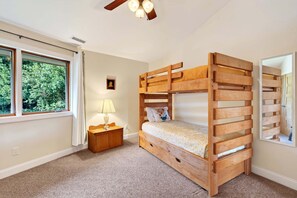 A bunk bed with a wooden frame, a bedside table with a lamp, a window with curtains, and a ceiling fan.
