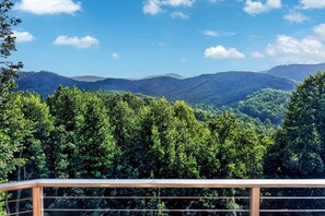 Vista desde un balcón con barandilla, que da a un bosque frondoso y montañas distantes bajo un cielo despejado.