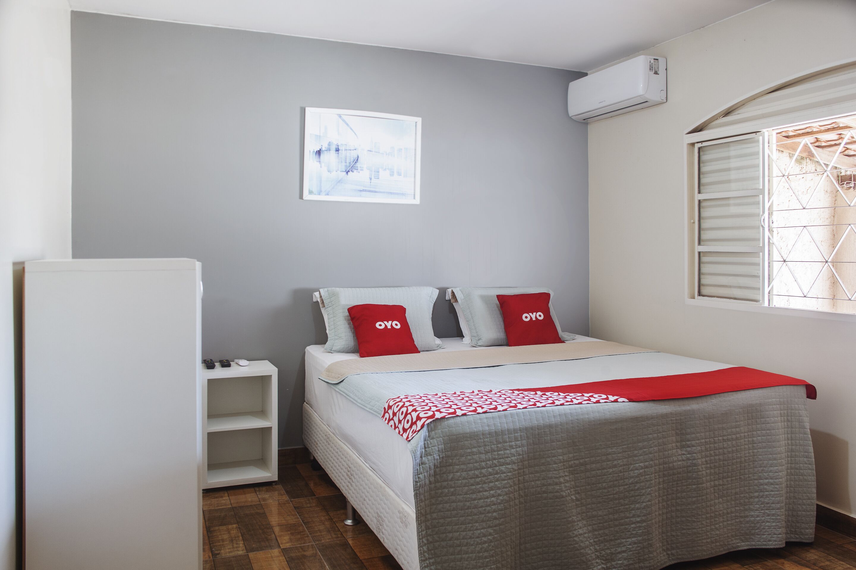 A bedroom with a bed, a white wardrobe, a small bedside table, a window with blinds, and a wall-mounted air conditioner.