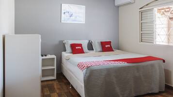 A bedroom with a bed, a white wardrobe, a small bedside table, a window with blinds, and a wall-mounted air conditioner.