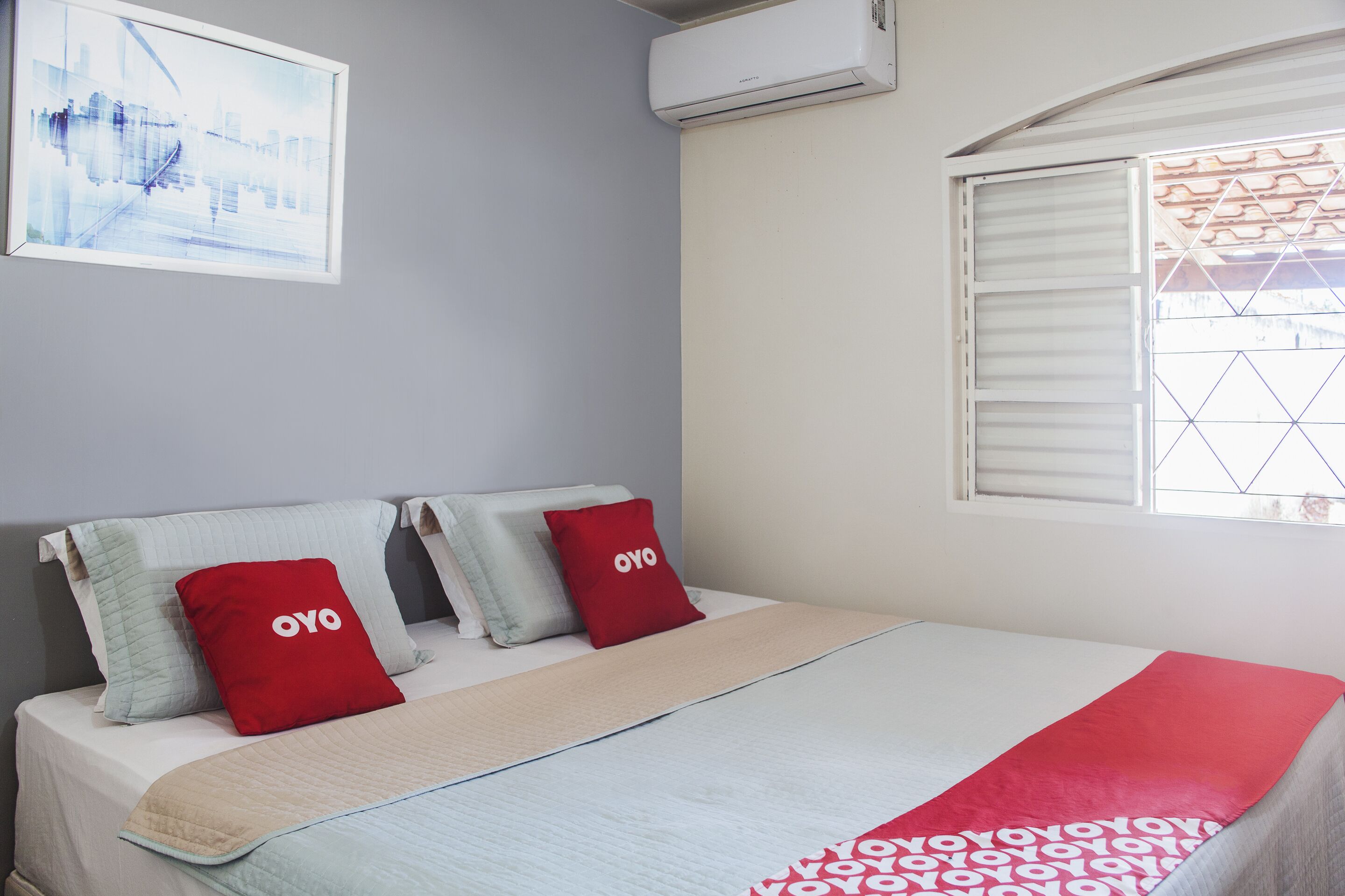 A neatly made bed with a red and white patterned blanket, two pillows with the word "OYO" on them, and a framed picture on the wall.