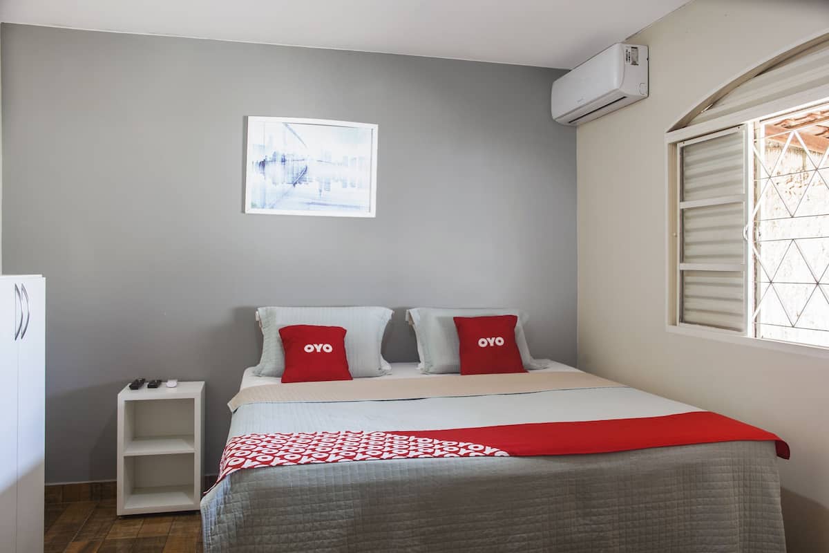 A bedroom with a bed, two pillows with 'OYO' branding, a bedside table, a wall-mounted air conditioner, and a window with blinds.