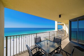 Un balcon avec une porte vitrée, une table en métal et des chaises, donnant sur une plage sous un ciel bleu dégagé.